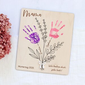 May include: Wooden sign with the word "Mama" at the top, two handprints in purple and pink, and the words "Muttertag 2025" and "Wir haben dich sehr lieb!" at the bottom.