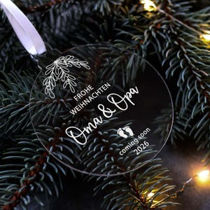 May include: A clear, round Christmas ornament with white text and a floral design. The ornament says "Frohe Weihnachten Oma & Opa coming soon 2026" and is hanging from a white ribbon. The background is a dark green Christmas tree.