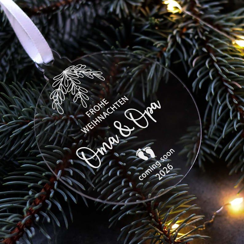 May include: A clear, round Christmas ornament with white text and a floral design. The ornament says "Frohe Weihnachten Oma & Opa coming soon 2026" and is hanging from a white ribbon. The background is a dark green Christmas tree.