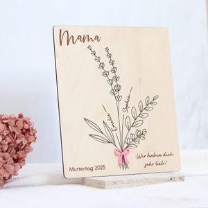 May include: Wooden sign with the word "Mama" at the top, a drawing of a bouquet of flowers with the names "Sophia" and "Emelie" written on the stems, and the text "Wir haben dich sehr lieb!" (We love you very much!) and "Muttertag 2025" (Mother's Day 2025) at the bottom.