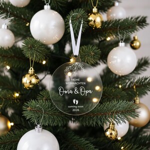 May include: A clear, round Christmas ornament with the text "Frohe Weihnachten Oma & Opa coming soon 2026" in white script. The ornament hangs from a white ribbon on a Christmas tree with white and gold ornaments.