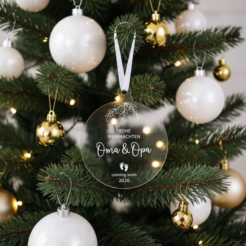 May include: A clear, round Christmas ornament with the text "Frohe Weihnachten Oma & Opa coming soon 2026" in white script. The ornament hangs from a white ribbon on a Christmas tree with white and gold ornaments.
