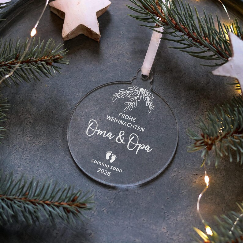 May include: A clear, round Christmas ornament with white text that reads "Frohe Weihnachten Oma & Opa coming soon 2026." The ornament is hanging by a white ribbon. The background includes a star, pine needles, and string lights.