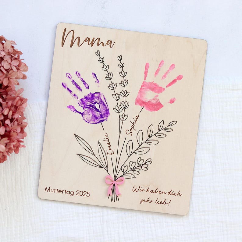 May include: Wooden sign with the word "Mama" at the top. Two handprints, one purple and one pink, are displayed as flowers with the names "Emeli" and "Sophia" written on the stems. The sign says "Muttertag 2025" and "Wir haben dich sehr lieb!"