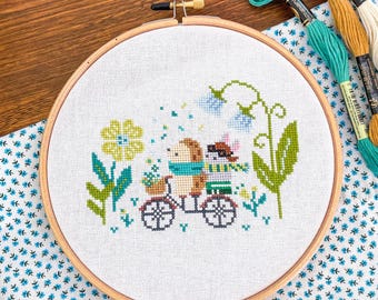 May include: Cross-stitch embroidery in a wooden hoop. The design features a hedgehog and a mouse riding a bicycle, surrounded by flowers. The color palette includes shades of green, blue, yellow, and brown. Embroidery floss in teal, gold, and white are visible.