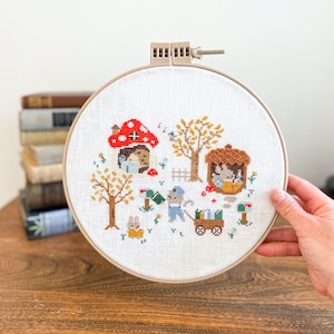 Könnte beinhalten: Ein Kreuzstich-Stickrahmen mit einer skurrilen Szene mit einem Pilzhaus, Bäumen und Figuren. Das Design umfasst ein Kaninchen, eine Maus, die einen Wagen zieht, und andere Elemente in Rot-, Braun-, Gelb- und Blautönen.