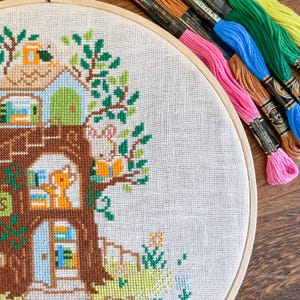 May include: A cross-stitch embroidery hoop featuring a whimsical treehouse scene with a rabbit reading a book. The design incorporates shades of brown, green, blue, and orange. Colorful embroidery floss in pink, blue, and green are visible.