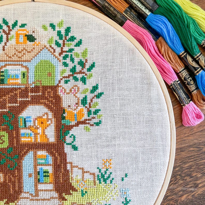 May include: A cross-stitch embroidery hoop featuring a whimsical treehouse scene with a rabbit reading a book. The design incorporates shades of brown, green, blue, and orange. Colorful embroidery floss in pink, blue, and green are visible.