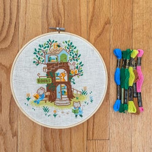 May include: A cross-stitch embroidery hoop featuring a whimsical treehouse scene with the word "BOOKS" on a sign. The scene includes animal characters reading. Colorful embroidery floss in blue, green, and pink is next to the hoop.