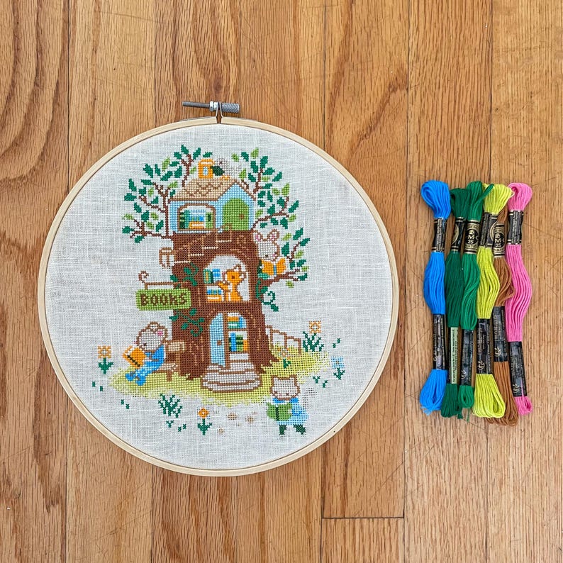 May include: A cross-stitch embroidery hoop featuring a whimsical treehouse scene with the word "BOOKS" on a sign. The scene includes animal characters reading. Colorful embroidery floss in blue, green, and pink is next to the hoop.