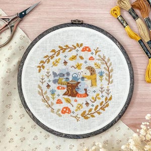 Può includere: Ricamo a punto croce completato in un telaio di legno. Il disegno presenta una scena boschiva con una figura centrale, circondata da una ghirlanda di foglie, ghiande e funghi. Fili da ricamo e forbici sono visibili.