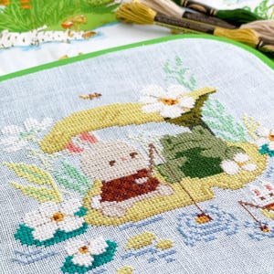May include: Cross-stitch embroidery of a bunny fishing on a lily pad, with a frog and flowers. The bunny wears a brown vest. The background is light blue with water and other aquatic plants. Embroidery floss is visible in the background.