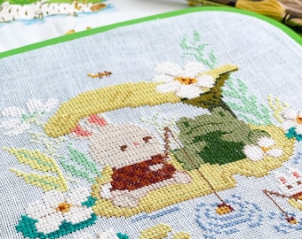 May include: Cross-stitch embroidery of a bunny fishing on a lily pad, with a frog and flowers. The bunny wears a brown vest. The background is light blue with water and other aquatic plants. Embroidery floss is visible in the background.