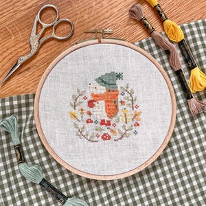Könnte beinhalten: Ein Kreuzstich-Stickrahmen mit einem herbstlichen Design, das einen Fuchs mit grünem Hut und floralem Kranz zeigt. Das Bild enthält gelbes, braunes und grünes Stickgarn sowie eine silberne Schere.