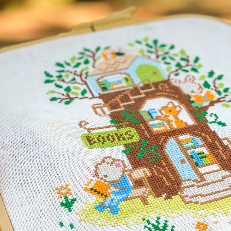 May include: Cross-stitch embroidery of a whimsical treehouse scene. The treehouse features a sign that reads "BOOKS", and is populated by various animal characters reading books. The color palette includes shades of brown, green, blue, and orange.