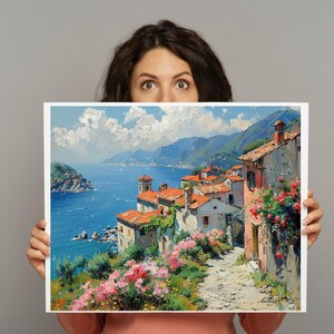 May include: An oil painting print depicting a coastal Mediterranean village with terracotta-roofed buildings, a stone path, and vibrant pink flowers overlooking a deep blue sea under a bright sky with white clouds, framed in a white border.