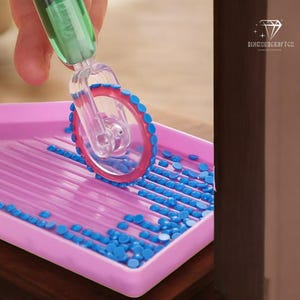 Puede incluir: Una bandeja de plástico rosa con cuentas azules en forma de diamante. Un rodillo de plástico transparente con un borde rojo está rodando sobre las cuentas.