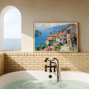 May include: A rectangular framed wall art print depicting a coastal Mediterranean village with terracotta-roofed buildings, vibrant pink flowers, and a deep blue sea under a bright sky, displayed on a light-colored brick wall above a white bathtub.