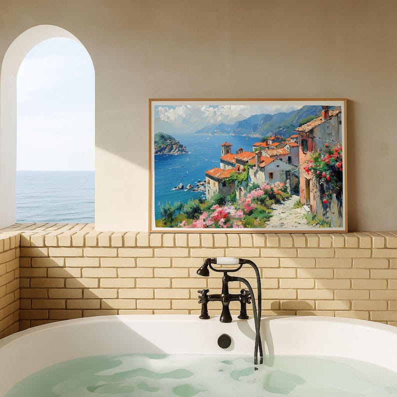 May include: A rectangular framed wall art print depicting a coastal Mediterranean village with terracotta-roofed buildings, vibrant pink flowers, and a deep blue sea under a bright sky, displayed on a light-colored brick wall above a white bathtub.