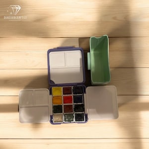 Peut inclure: Un coffret de peinture aquarelle violet et blanc avec 12 godets de peinture, un porte-pinceaux et un couvercle. Le coffret de peinture est ouvert et les godets de peinture sont visibles. Le coffret de peinture est sur une surface en bois.