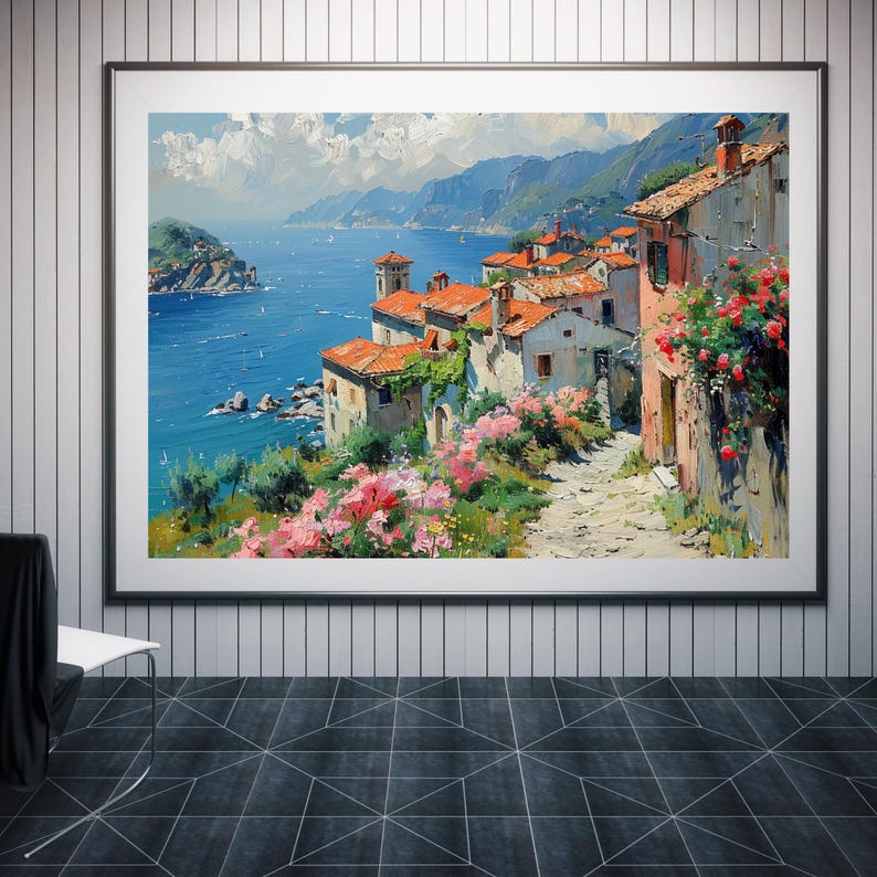 May include: A framed landscape painting depicting a coastal village with terracotta-roofed buildings, stone pathways, and vibrant pink flowers overlooking a deep blue sea under a bright sky with white clouds, all set within a simple black frame.