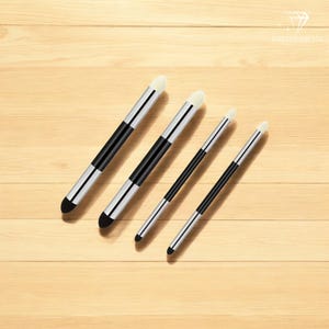 Pode incluir: Quatro pincéis de maquiagem com cabos pretos e prateados e cerdas brancas. Os pincéis estão organizados por tamanho, com o pincel maior à esquerda e o menor à direita.