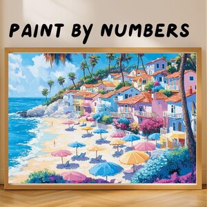 May include: A framed "Paint by Numbers" painting depicts a vibrant beach scene. Colorful umbrellas dot the sandy shore, with buildings nestled on a hillside. The artwork features a bright blue sky, turquoise water, and lush green palm trees.