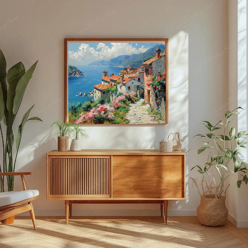 May include: A rectangular framed wall art print depicting a coastal Mediterranean village scene with terracotta-roofed buildings, a stone path, vibrant pink flowers, and a deep blue sea under a bright sky with white clouds, mounted on a wall.