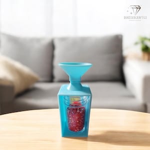 Puede incluir: Un molinillo de especias azul claro con una parte superior en forma de embudo y un recipiente transparente lleno de granos de pimienta roja. El molinillo es de plástico y está sobre una mesa de madera. El fondo está borroso, mostrando un sofá.