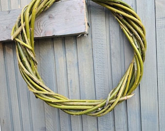 May include: A circular wreath crafted from woven green willow branches. The wreath is displayed on a grey wooden fence. The branches are various shades of green, giving a natural, rustic appearance.