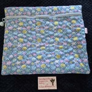 May include: A quilted pouch with a zip closure, decorated with a sea-themed pattern. The design includes seashells and starfish in pastel shades of yellow, pink, and blue on a light blue backdrop. The pouch has a light blue zip and a small pom-pom.