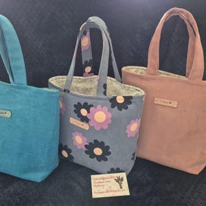May include: Three corduroy tote bags in teal, floral print, and dusky pink. Each bag has a matching handle and a small rectangular tag. The floral bag has a grey background with pink and black flowers. The bags are lined with a patterned fabric.