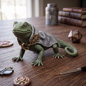 May include: A green lizard figurine wearing a grey cape and a gold-colored metal chain with a crescent moon charm. The lizard has textured skin and is positioned on a wooden surface alongside a twenty-sided die, small tokens, and a paintbrush.