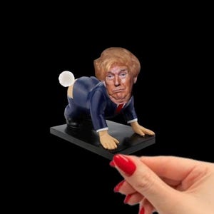 Könnte beinhalten: Ein Spielzeug aus dem Bereich der Neuheiten, das eine Person in einem blauen Anzug mit weißem Hemd und roter Krawatte darstellt, mit einem Gesicht, das Donald Trump ähnelt, auf allen Vieren kriechend mit einem weißen Ball, der an seinem Hinterteil befestigt ist.