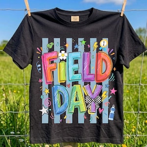 Op de afbeelding: Zwart t-shirt met de tekst "FIELD DAY" in kleurrijke, afgeronde letters. Het ontwerp bevat sterren, bliksemschichten en een geblokte vlag. Het t-shirt hangt aan een waslijn voor een groen veld.
