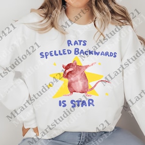 May include: White sweatshirt with a graphic featuring a pink rat dabbing on a yellow star. The text reads "RATS SPELLED BACKWARDS IS STAR" in blue. The design includes small yellow stars.