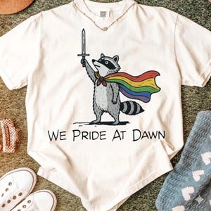 Op de afbeelding: Crèmekleurig T-shirt met een cartoon wasbeer met een regenboogcape en een zwaard. De tekst "WE PRIDE AT DAWN" staat eronder. Een zilveren ketting is zichtbaar.