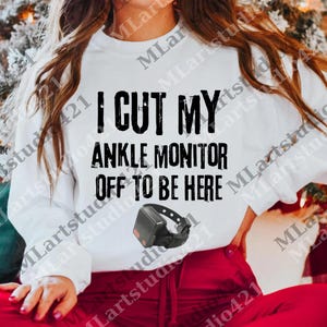 Könnte beinhalten: Weißes Sweatshirt mit dem Text "I CUT MY ANKLE MONITOR OFF TO BE HERE" in schwarzen Blockbuchstaben. Unter dem Text ist eine schwarze Fußfessel abgebildet. Das Sweatshirt wird mit einer roten Hose kombiniert.