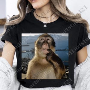 Puede incluir: Camiseta negra con una imagen de un mono pensativo, con el dedo en la boca. El mono tiene pelaje marrón y beige. El fondo muestra una masa de agua y una costa distante.