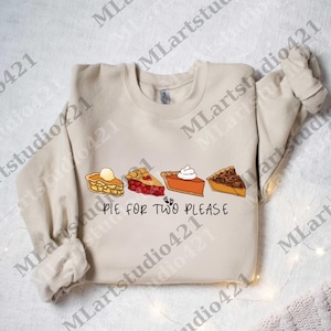 Puede incluir: Sudadera beige con un gráfico de cuatro porciones de pastel: manzana, cereza, calabaza y nuez. El texto "PIE FOR TWO PLEASE" está impreso debajo. La sudadera tiene mangas largas y cuello redondo.