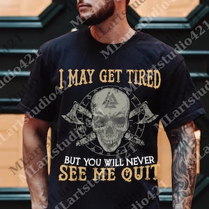 May include: Black t-shirt with a skull graphic, crossed axes, and the text "I MAY GET TIRED BUT YOU WILL NEVER SEE ME QUIT" in gold lettering. The design includes a Viking symbol.