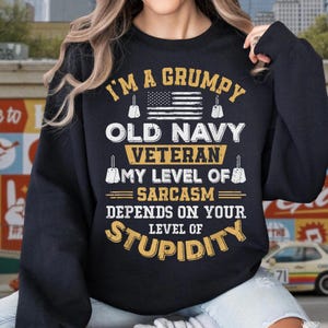May include: A black long-sleeve sweatshirt featuring yellow and white text that reads I'M A GRUMPY OLD NAVY VETERAN MY LEVEL OF SARCASM DEPENDS ON YOUR LEVEL OF STUPIDITY. The design includes a graphic of an American flag and four dog tags.