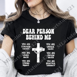 May include: Black t-shirt with white text and a cross graphic. The text reads "DEAR PERSON BEHIND ME" with inspirational messages like "YOU ARE FORGIVEN" and "YOU ARE UNIQUE".