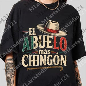 Può includere: T-shirt nero con un design grafico con la scritta "EL ABUELO más CHINGÓN" in rosso, verde e beige. Un cappello con una fascia rossa è sopra il testo. Un bastone è sotto.