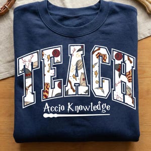 May include: A navy blue crewneck sweatshirt featuring the word TEACH in large white block letters filled with a pattern of magical icons including wands, hats, and golden snitches. Below, the text Accio Knowledge appears in white script.