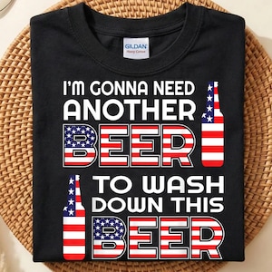 May include: A black short-sleeve t-shirt featuring the white text I'M GONNA NEED ANOTHER BEER TO WASH DOWN THIS BEER. The words BEER are filled with a red, white, and blue American flag pattern, accompanied by two bottle graphics with the same flag.