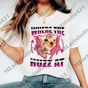 Op de afbeelding: Wit T-shirt met een afbeelding van een katje in een roze bliksemontwerp. De tekst "WHERE THE HUZZ AT" staat boven en onder de kattenafbeelding in een paarse en zwarte lettertype.