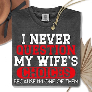 Op de afbeelding: Donkergrijs t-shirt met de tekst "I NEVER QUESTION MY WIFE'S CHOICES BECAUSE I'M ONE OF THEM" in wit en rood. Een gouden ketting met een hartvormige hanger is over het shirt gedrapeerd.