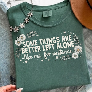May include: A forest green short-sleeve t-shirt featuring the white text SOME THINGS ARE BETTER LEFT ALONE like me, for instance, surrounded by white daisy illustrations, small stars, and hearts, with a silver floral necklace draped across the top.