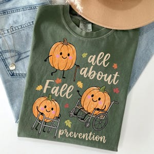 May include: An olive green t-shirt featuring a graphic of three orange pumpkins with faces. One pumpkin uses a cane, one uses a walker, and one sits in a wheelchair. The text reads "all about Fall prevention" in a cursive font, surrounded by small autumn leaves.